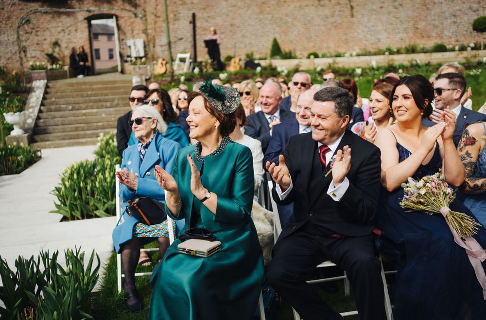Wedding guests applauding during an outdoor ceremony with brick wall and garden setting.