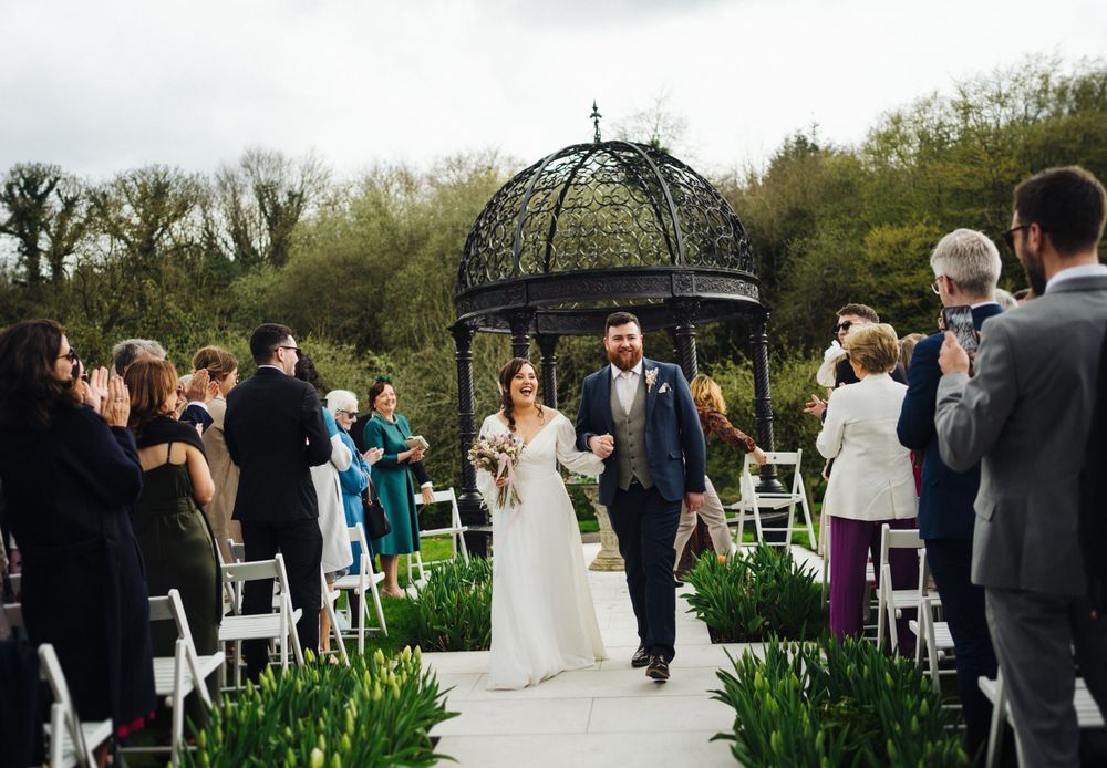 Newlyweds walking down the aisle under a black metal gazebo surrounded by spring flowers.