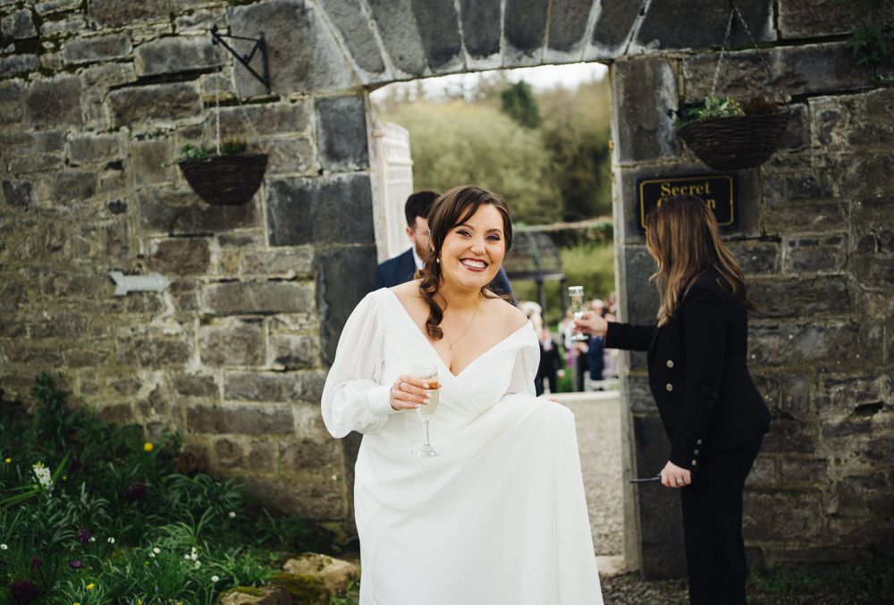 A bride in a white dress stands smiling in front of a rustic stone archway during an outdoor wedding celebration.