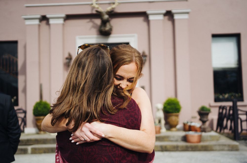 Two people share a warm embrace outside a pink-colored building with decorative columns.