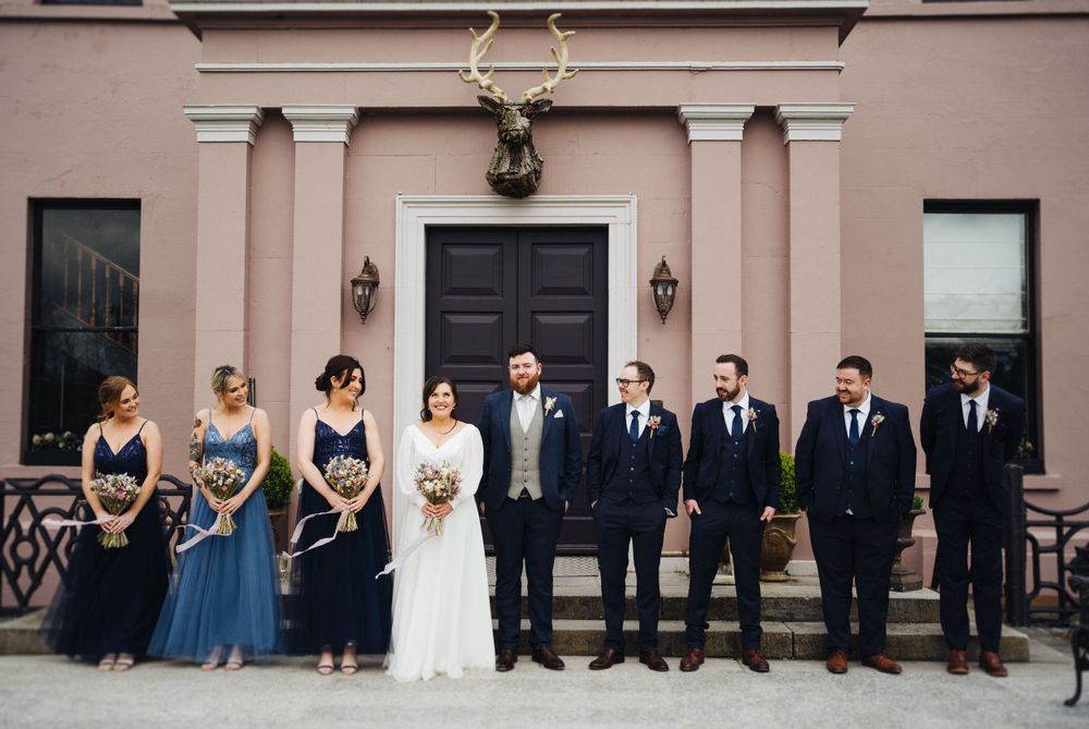 Wedding party poses formally outside pink building with bridal party in navy and white attire.