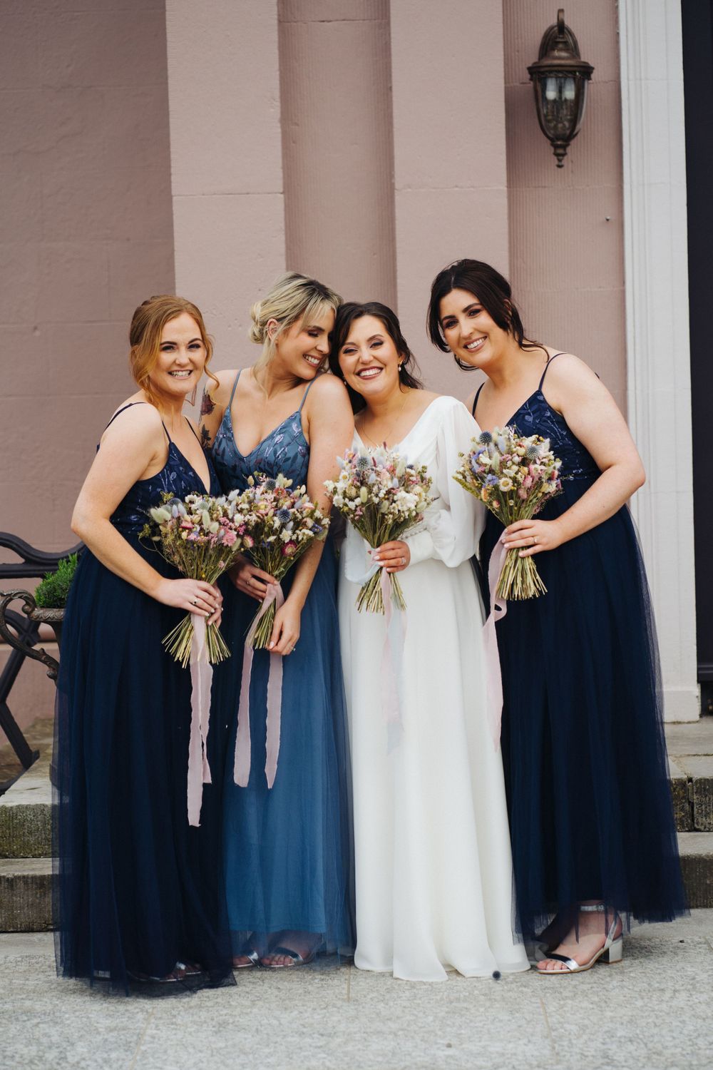 Bridesmaids in navy blue dresses pose with bouquets on building steps.