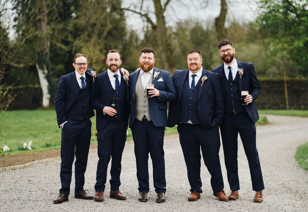 Five groomsmen in navy suits pose casually on outdoor path.