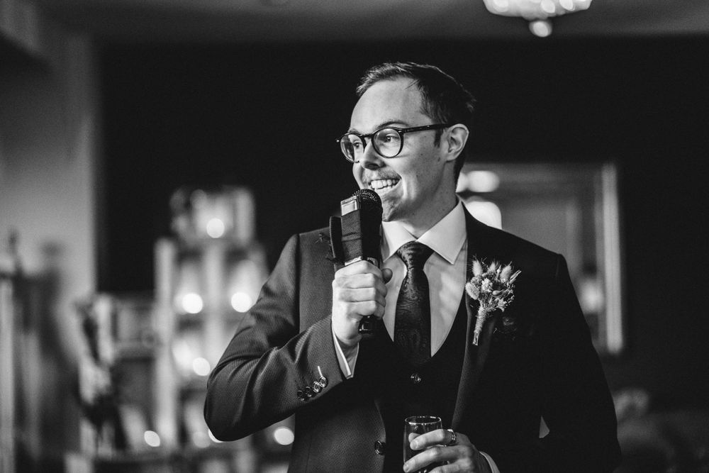 A speaker in a suit and glasses holds a microphone while giving a wedding speech at an indoor reception.