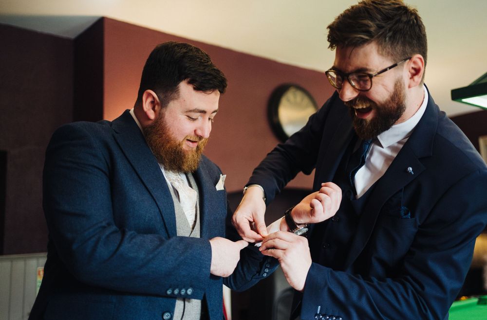 Two groomsmen in navy suits share a lighthearted moment while getting ready for the wedding ceremony.