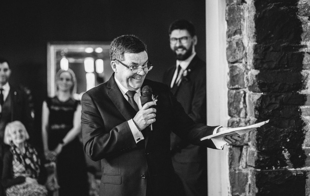 A speaker delivers remarks at a wedding reception while holding notes and a microphone.