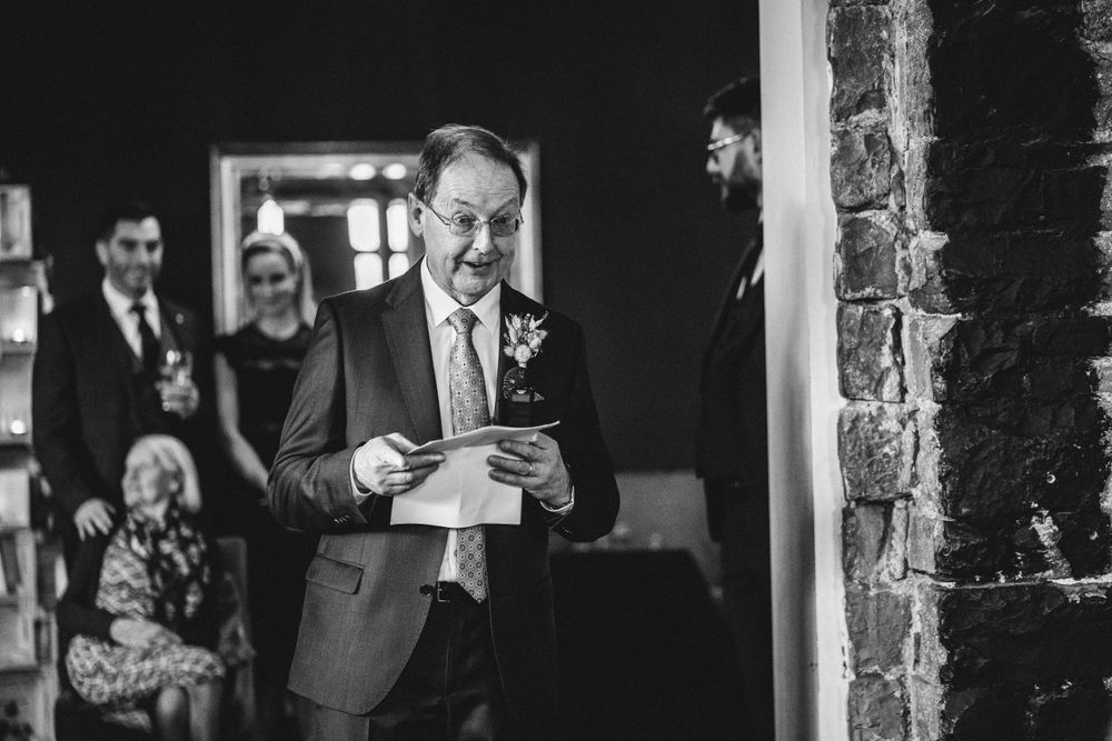 A speaker in a dark suit reads from papers during a wedding speech.