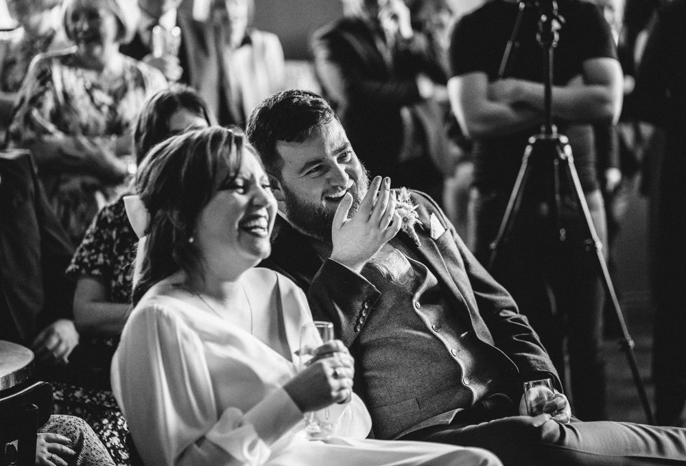 Guests laugh while enjoying speeches during an indoor wedding celebration.