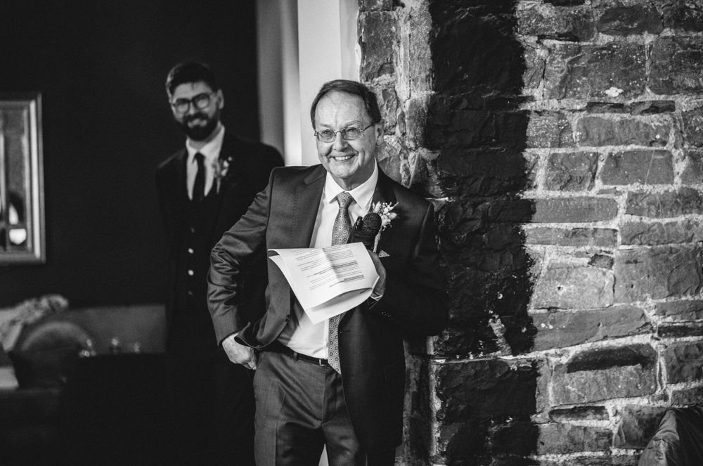 A speaker smiles while holding papers during a wedding speech near exposed brick walls.