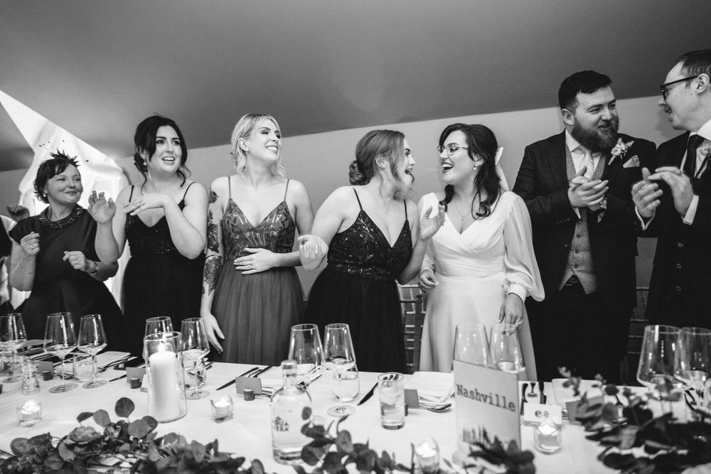 Wedding party laughs together while standing at decorated reception table.