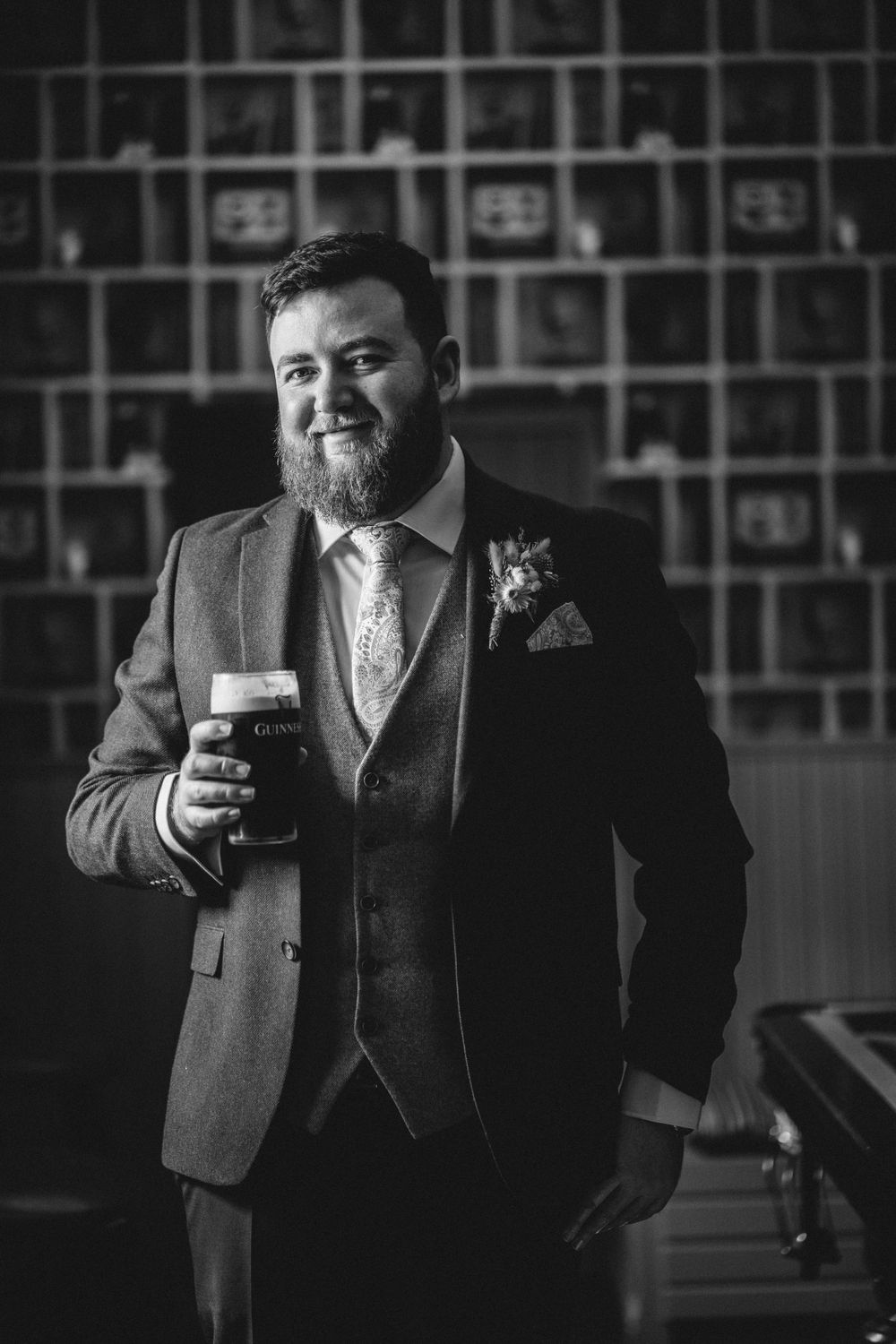 Groom in grey three-piece suit holds a dark beer while posing against geometric wall pattern.