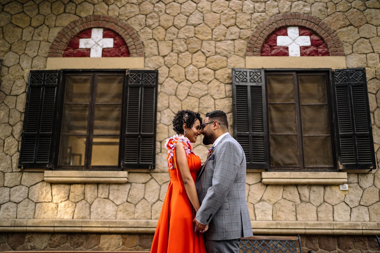 Couple in formal attire stands close together against a stone wall with cross-decorated windows.
