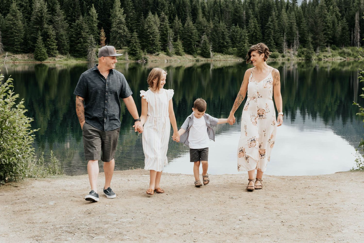A family of four takes a stroll along a serene mountain lake path surrounded by evergreen forest.