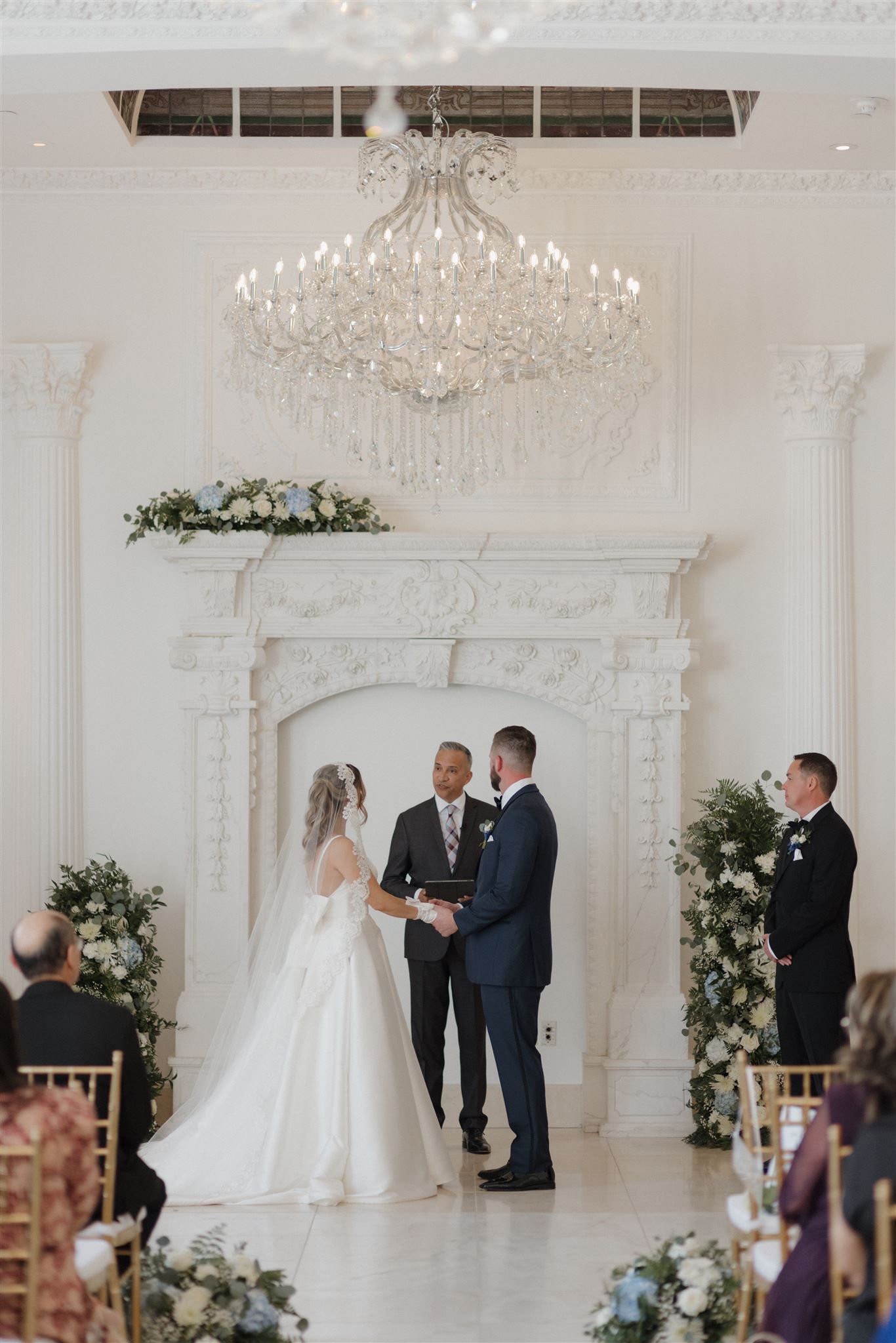 An elegant wedding ceremony with ornate chandelier and floral arrangements in a white room.