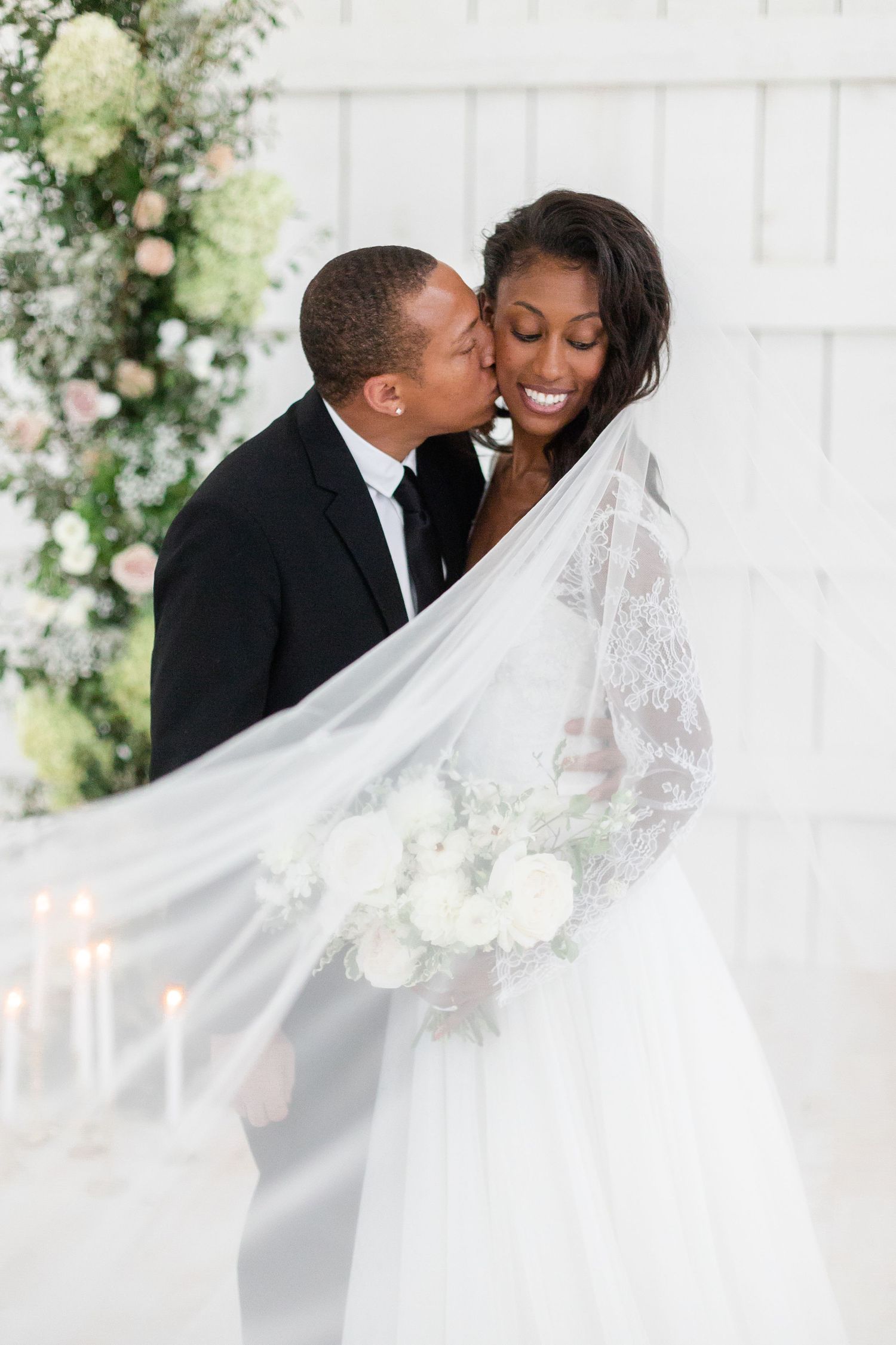 Elegant wedding couple embraces against white backdrop with flowing lace veil and floral decorations.