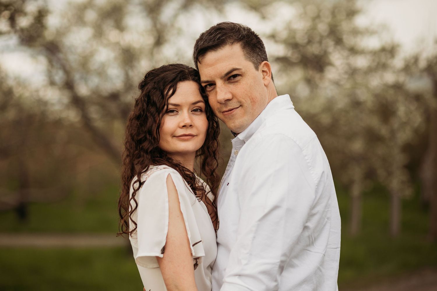 A romantic couple poses together in a spring orchard with blooming trees in the background.