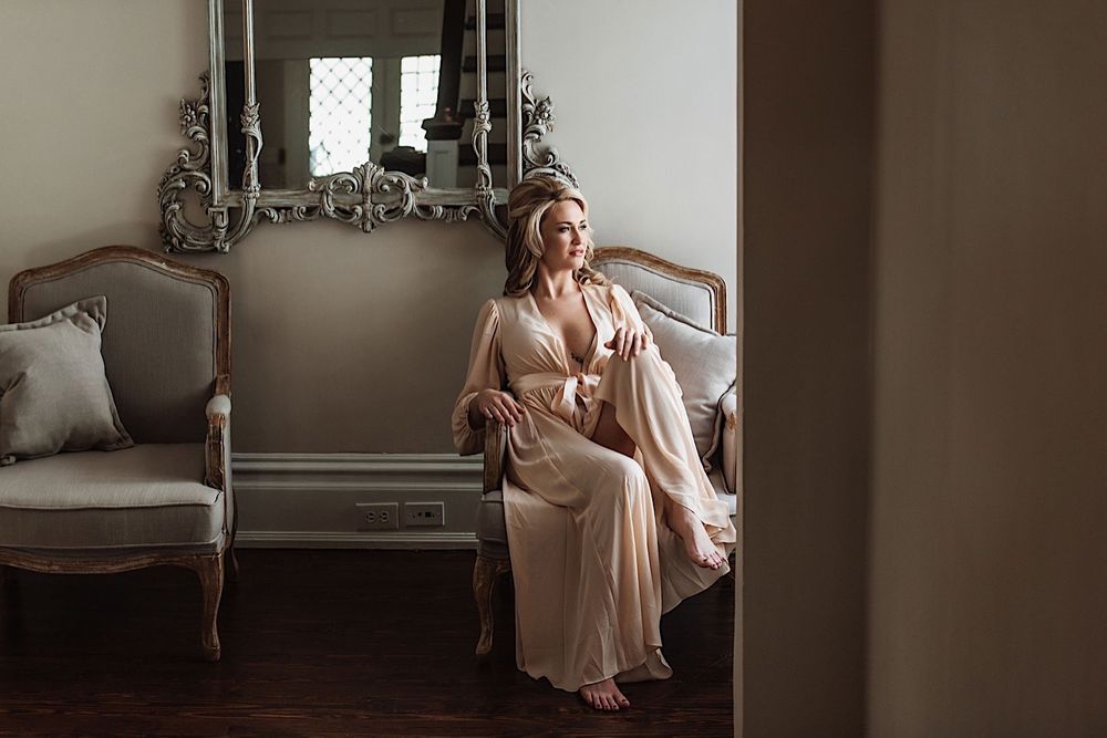 Bride sitting on a love seat in peach silk robe and looking out the window