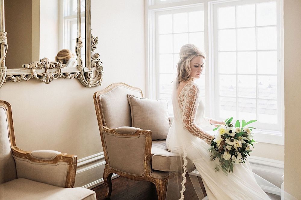 Bride in wedding dress with bouquet sitting on  chair in front of the window at The Orlo