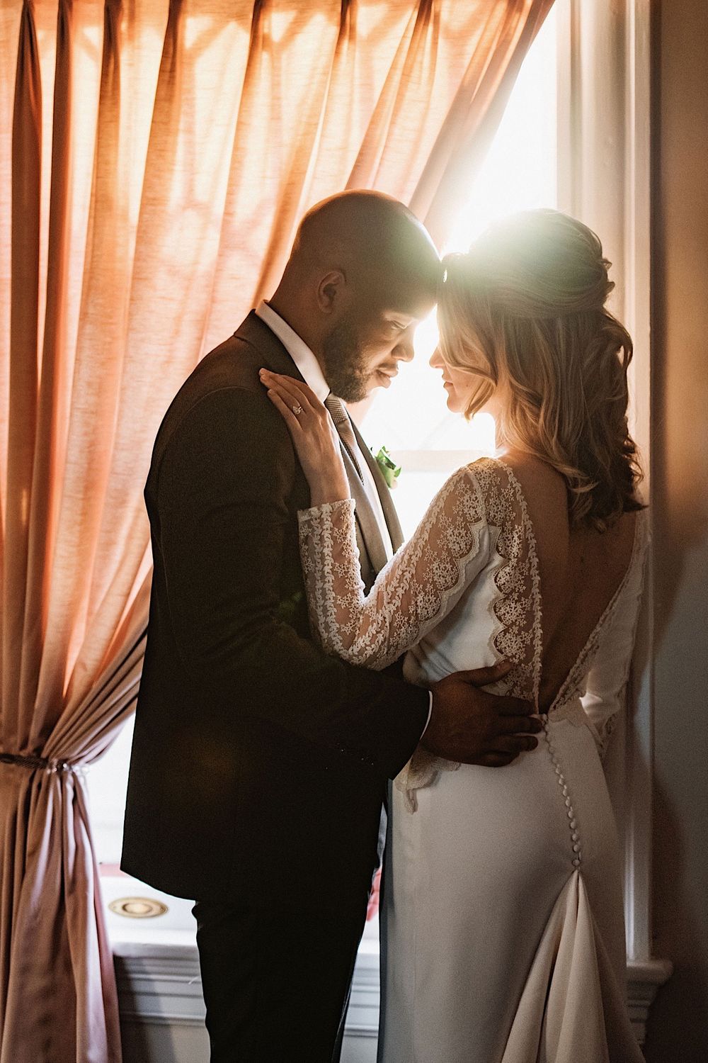 bride and groom touching foreheads in front of window with pink curtain at the orlo