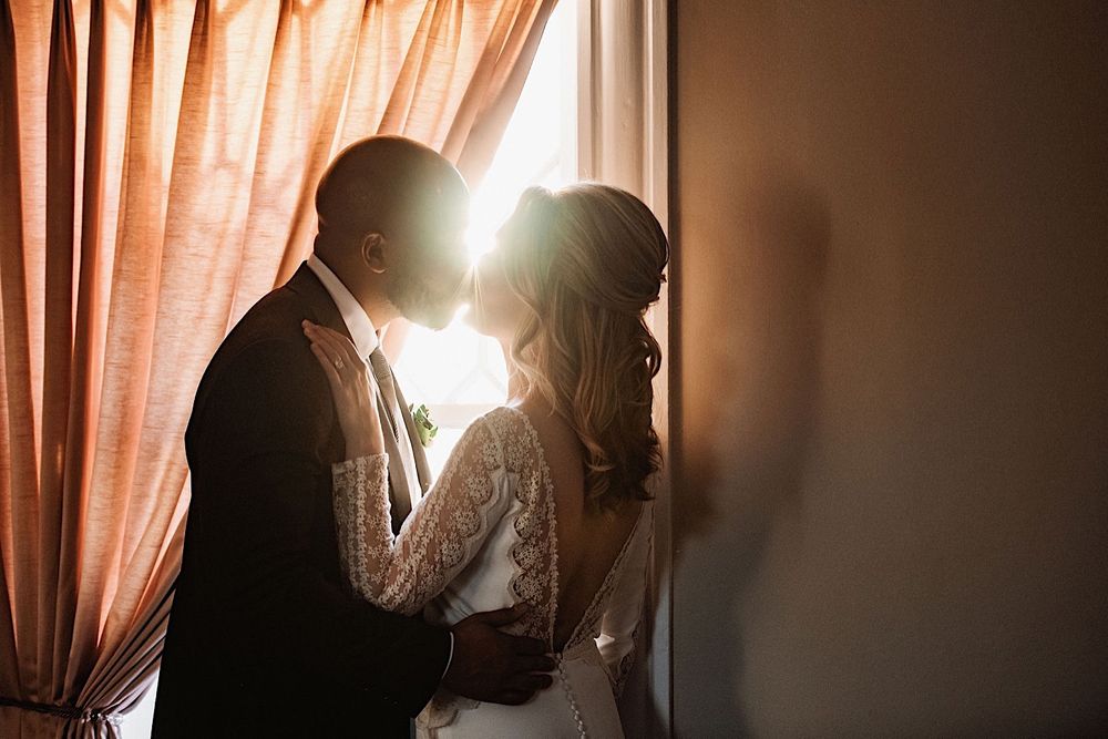 bride and groom kissing in front of a window with sunlight shining thorugh
