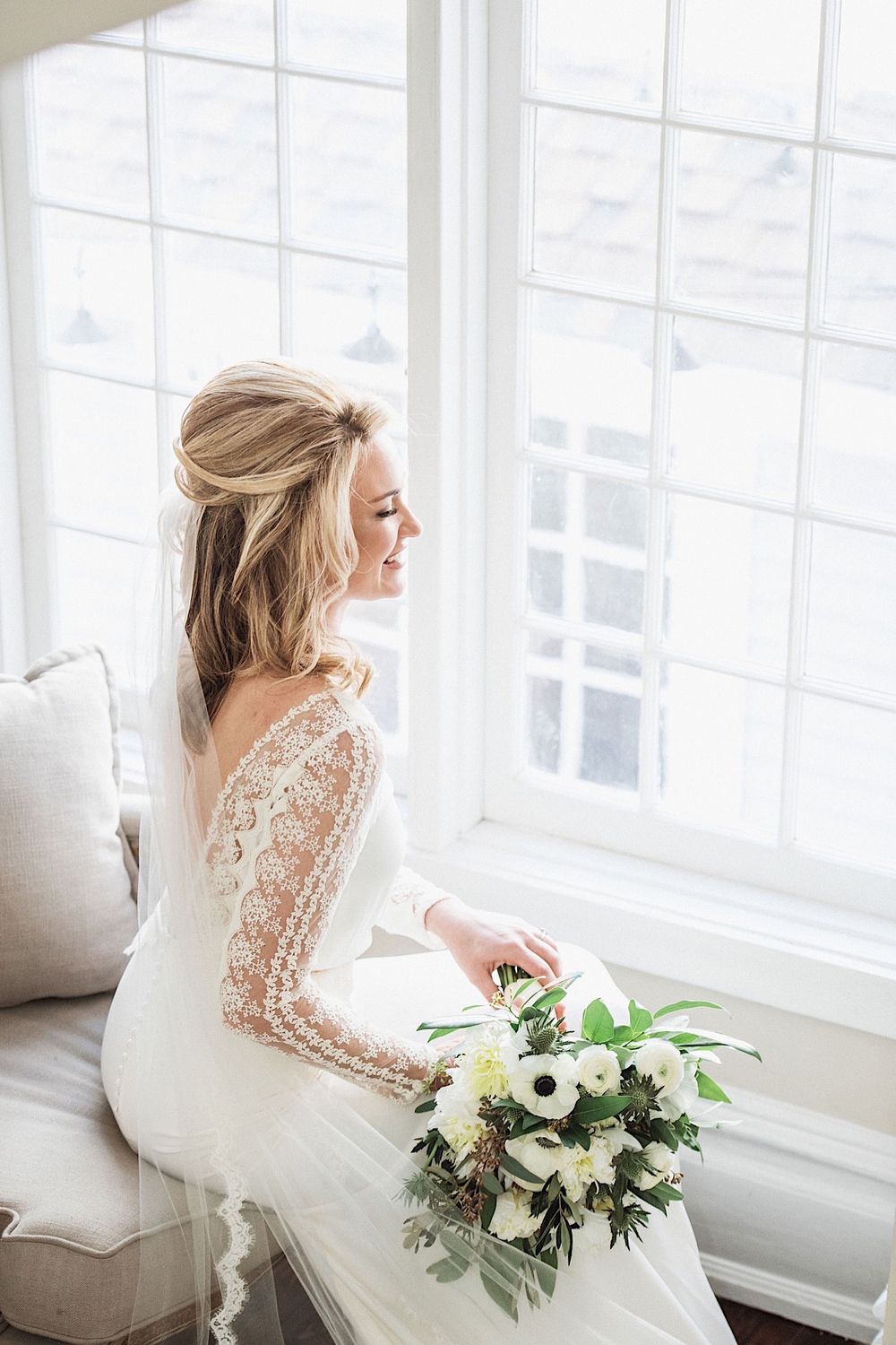 bride looking out the window while holding bouquet and wearing a BHLDN gown