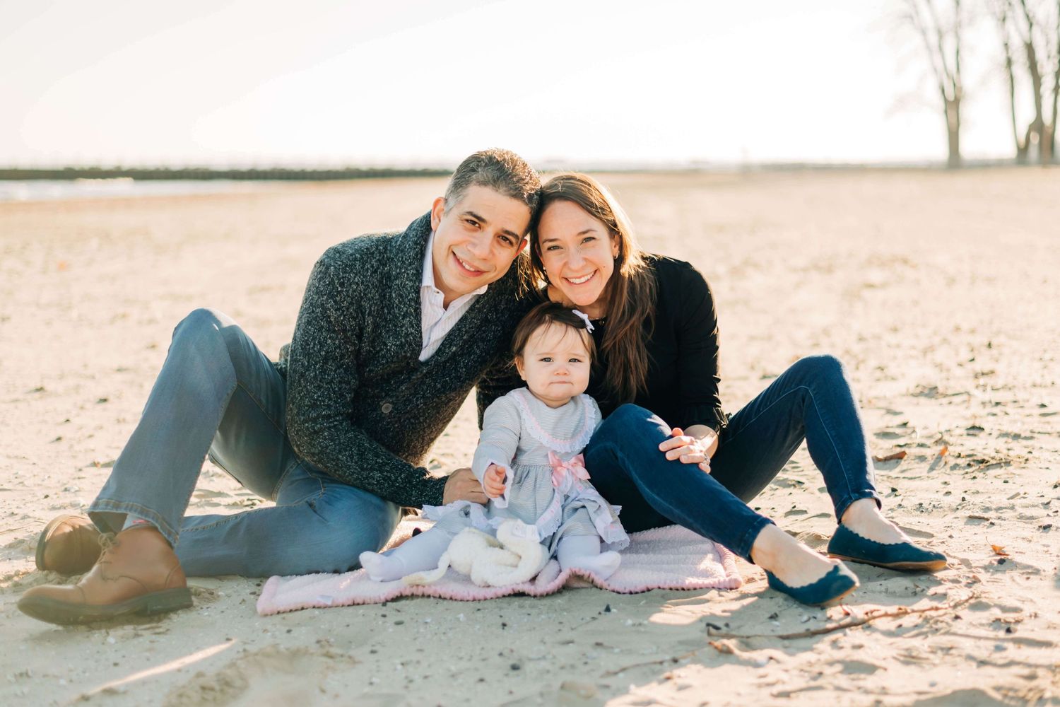 Photo of mother, father, and baby girl sitting in the sand at a beach