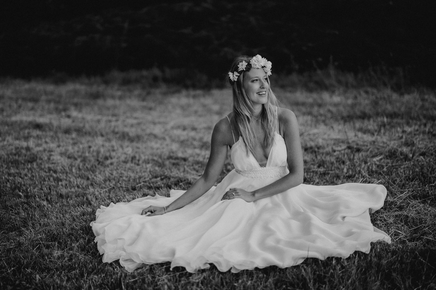 Ethereal black and white portrait of bride in flowing dress sitting in field wearing flower crown.