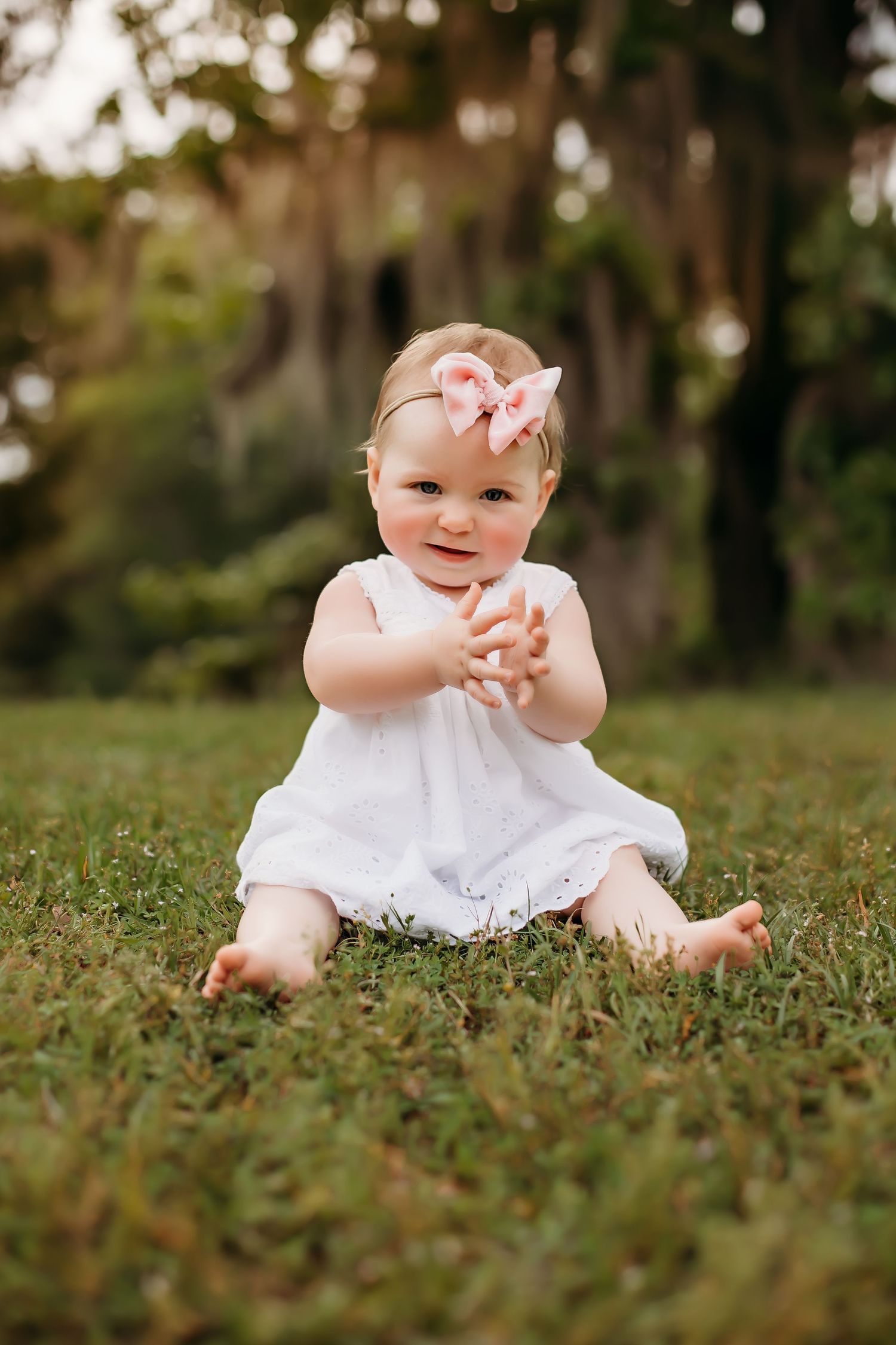 Baby in white dress sitting on grass wearing pink flower hair accessory.