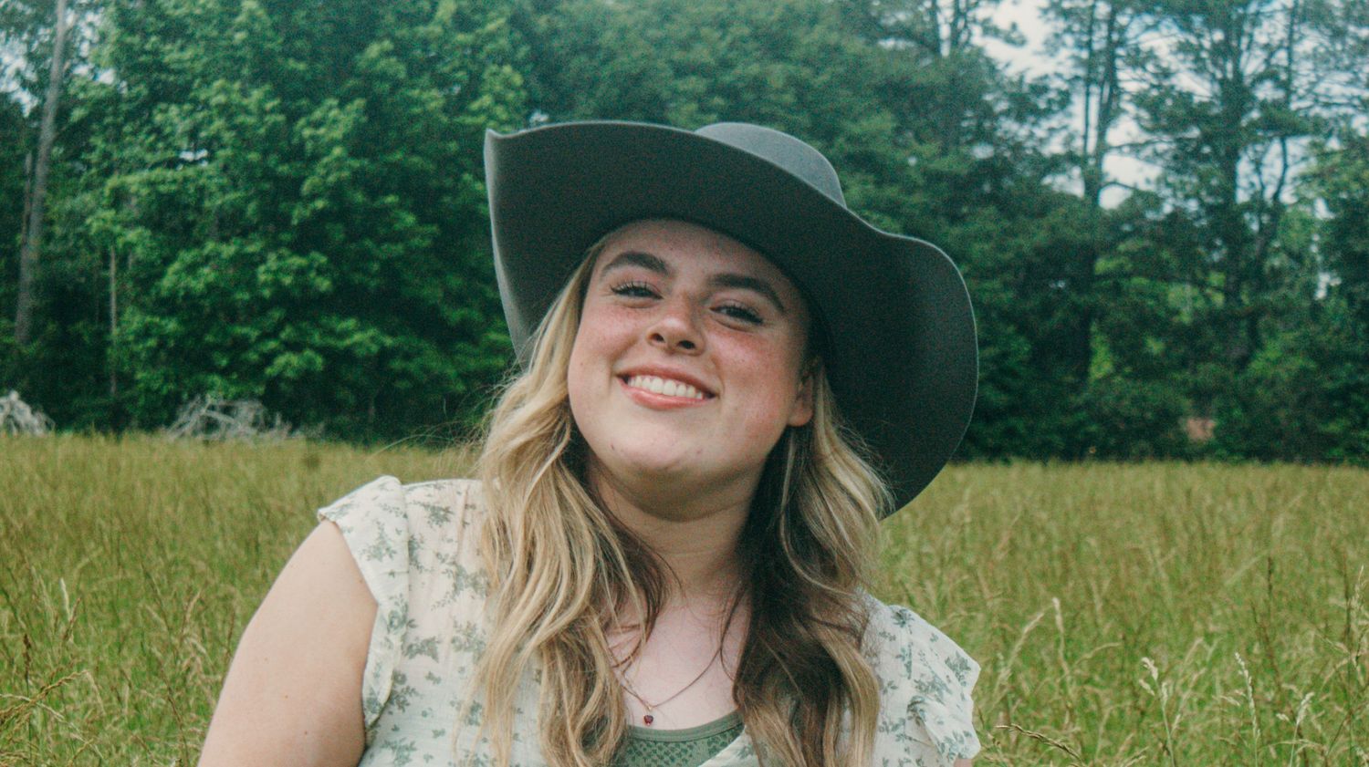 A person wearing a wide-brimmed hat smiles in a sunny meadow with trees in background.