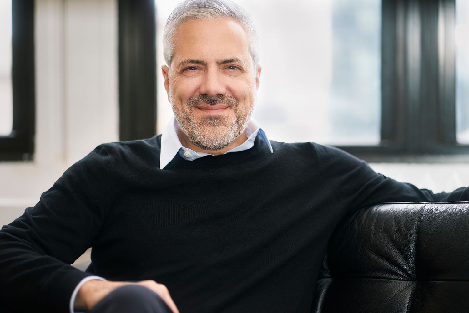 Smiling middle-aged man with gray hair and beard wearing a black sweater, relaxing on a leather sofa.