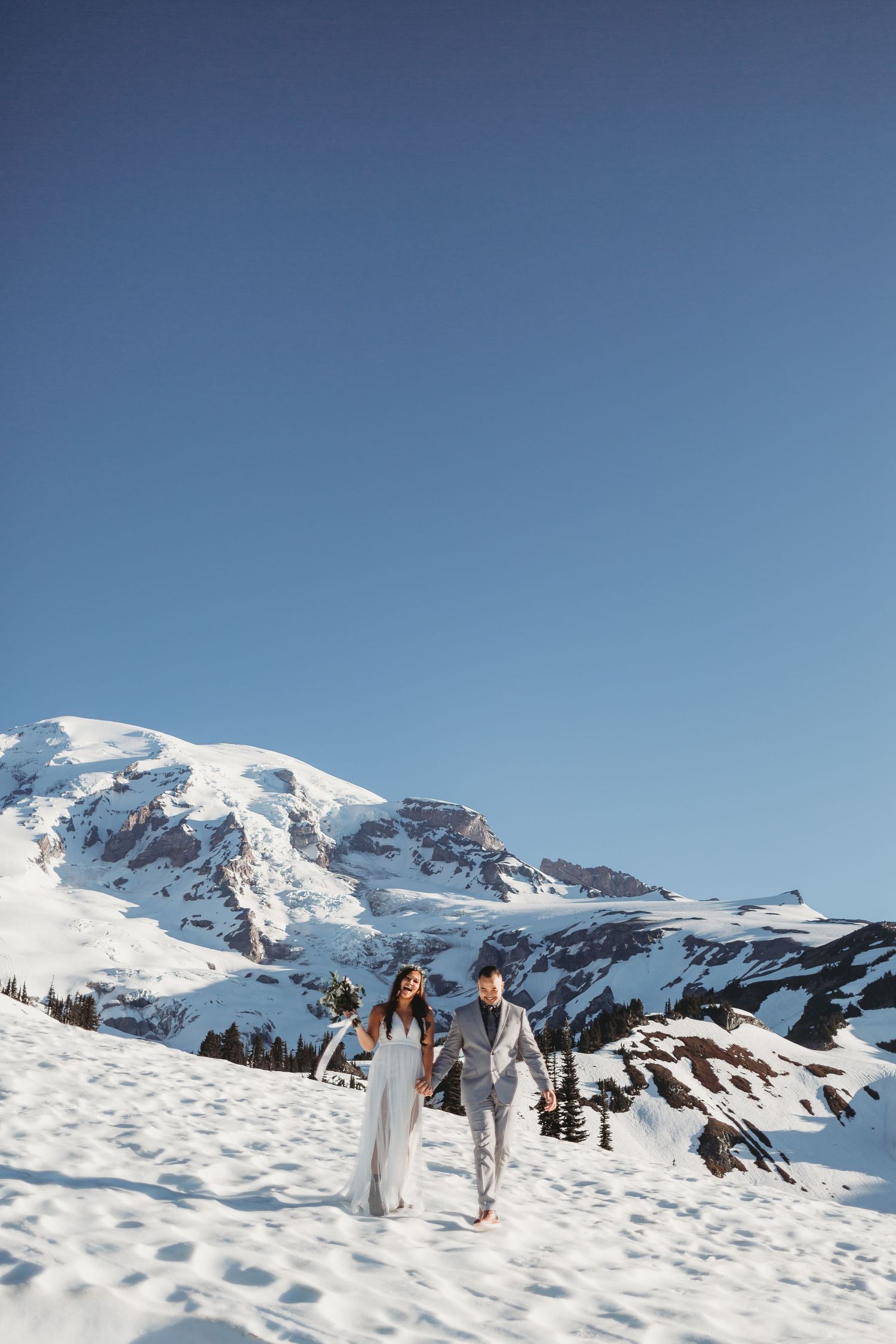 A laughing bride and groom walk down the snow bank of a hill with Mount Rainier just behind them at their vow renewal.