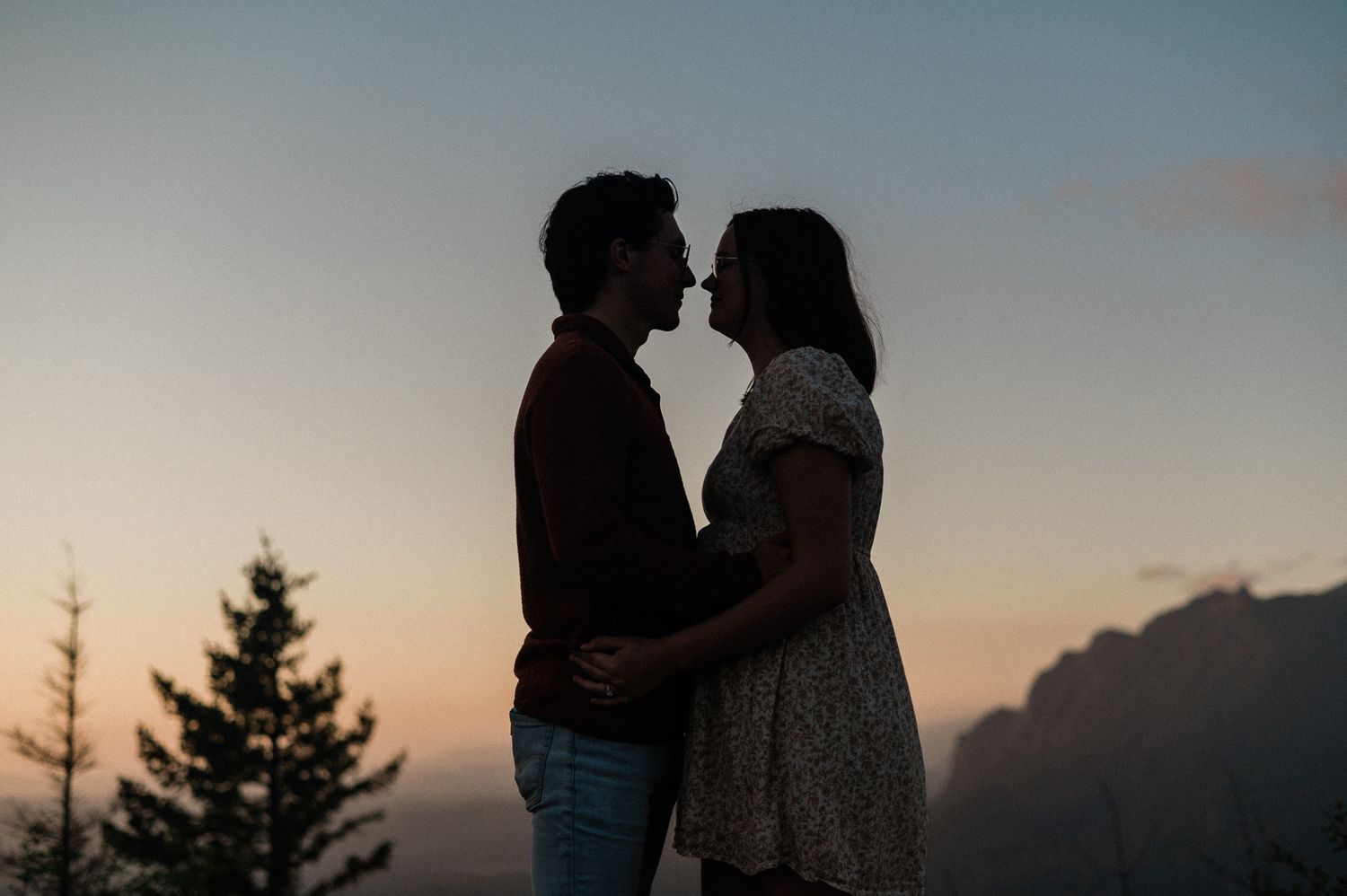 Silhouetted couple embracing against a sunset sky with evergreen trees in the background.