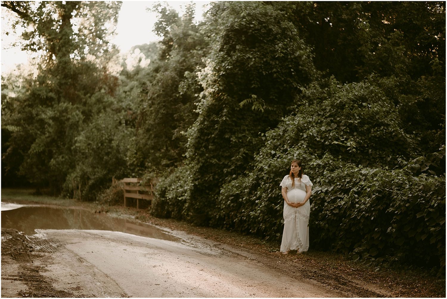 Distant view of expectant mother in white dress standing on curved forest path.