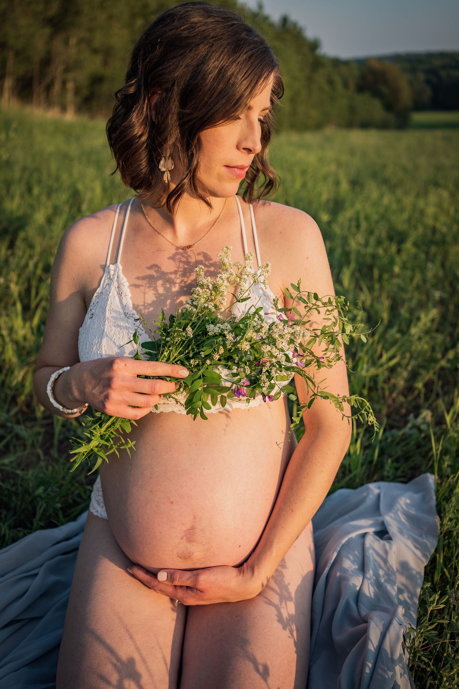 Expectant mother in white dress holds greenery while standing in a sunny meadow during maternity photoshoot.