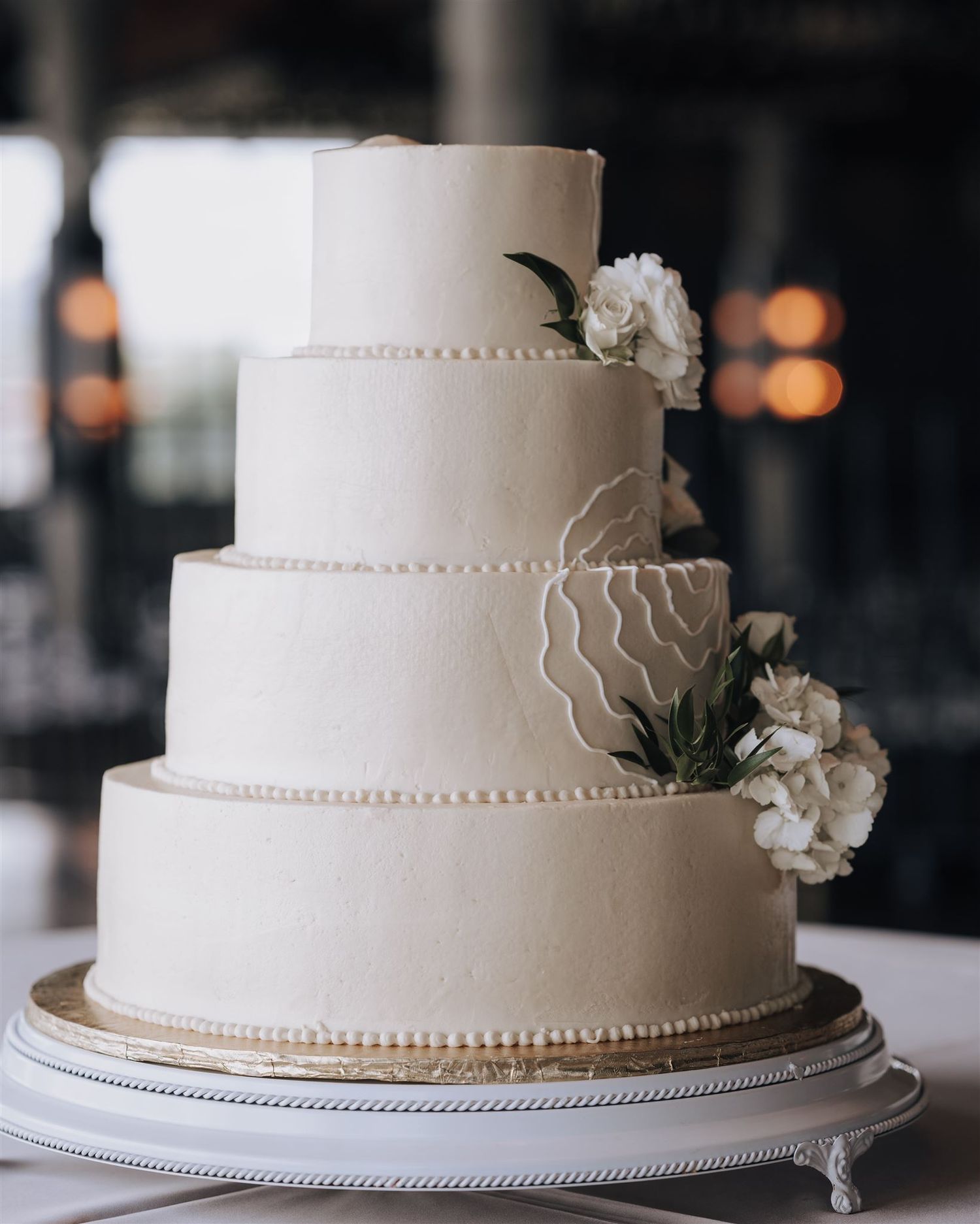 Elegant four-tier white wedding cake with pearl details and white floral decorations on a silver stand.