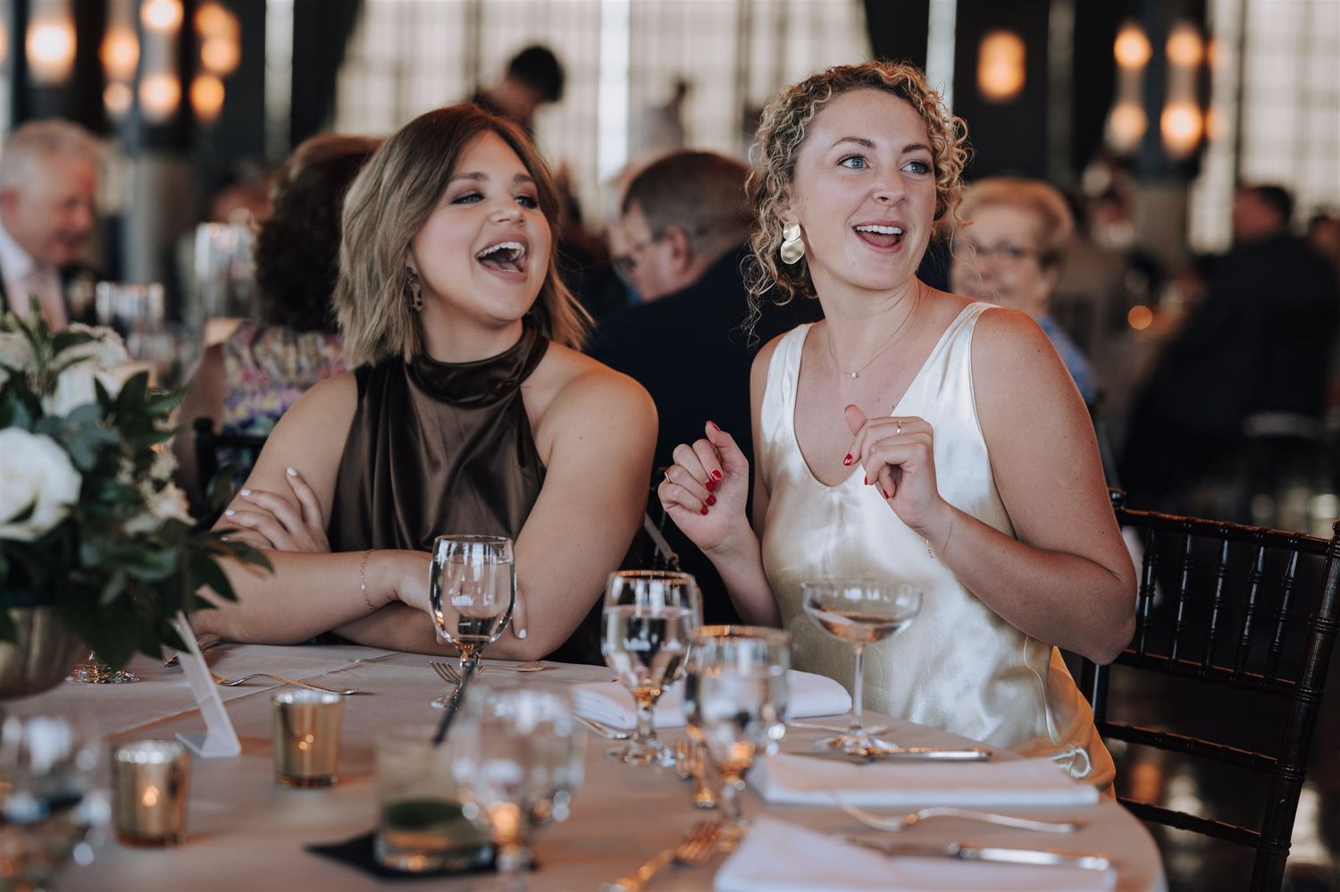 Two women laugh joyfully at an elegant reception dinner with wine glasses and candlelight.