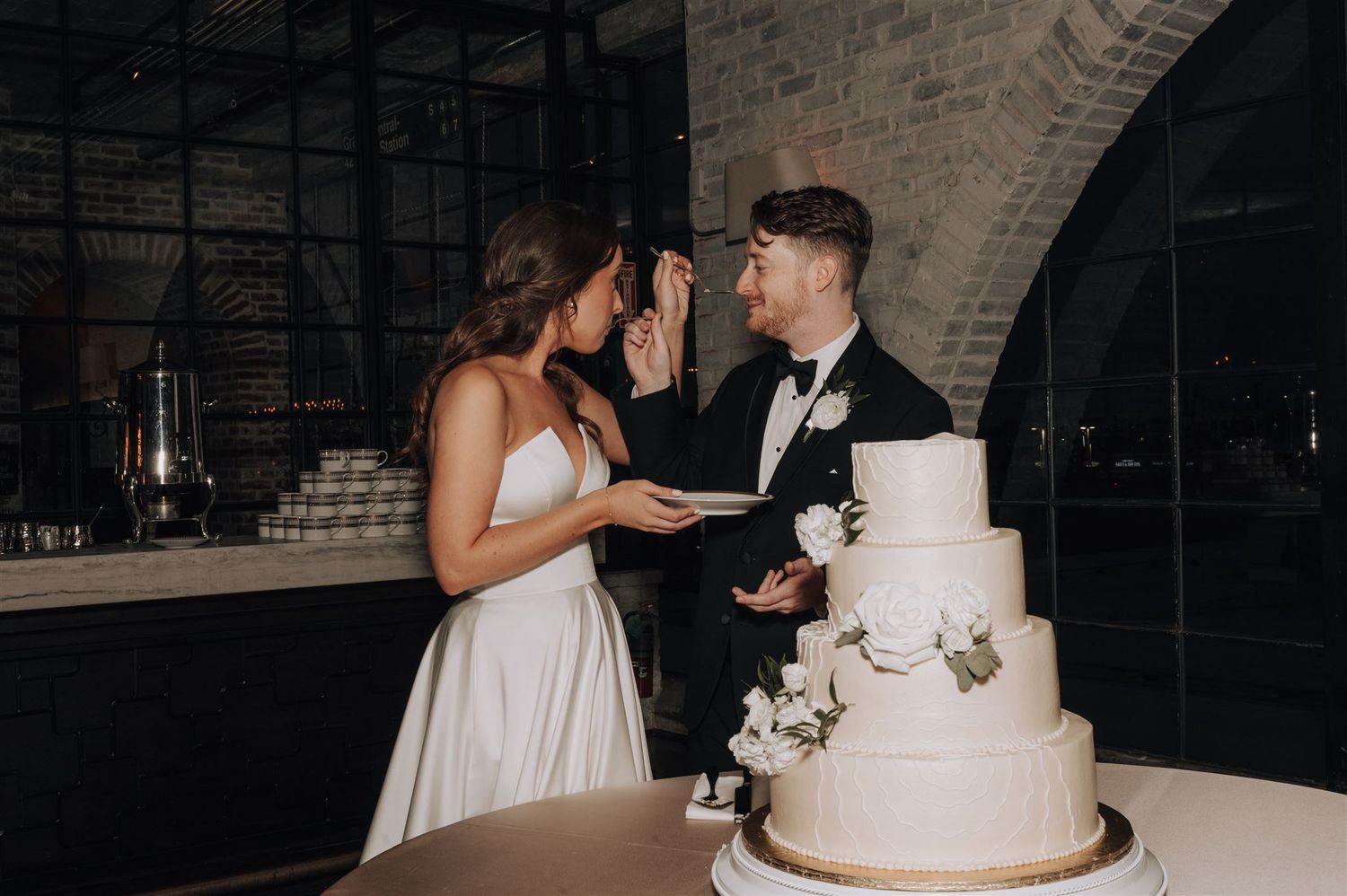 Wedding couple sharing cake cutting moment at reception with elegant white tiered cake.