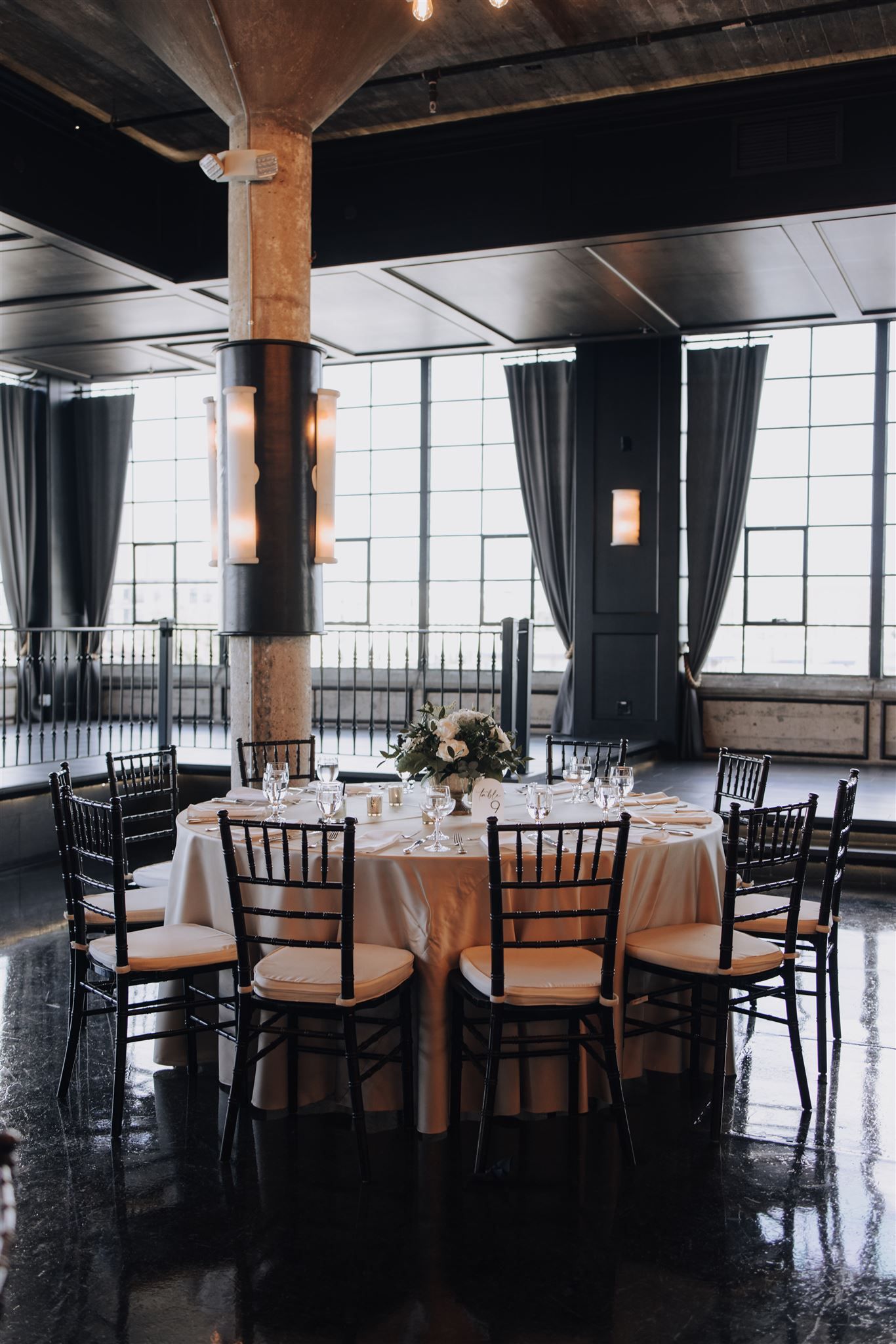 Modern industrial wedding venue with large windows, dark curtains, and black chiavari chairs.