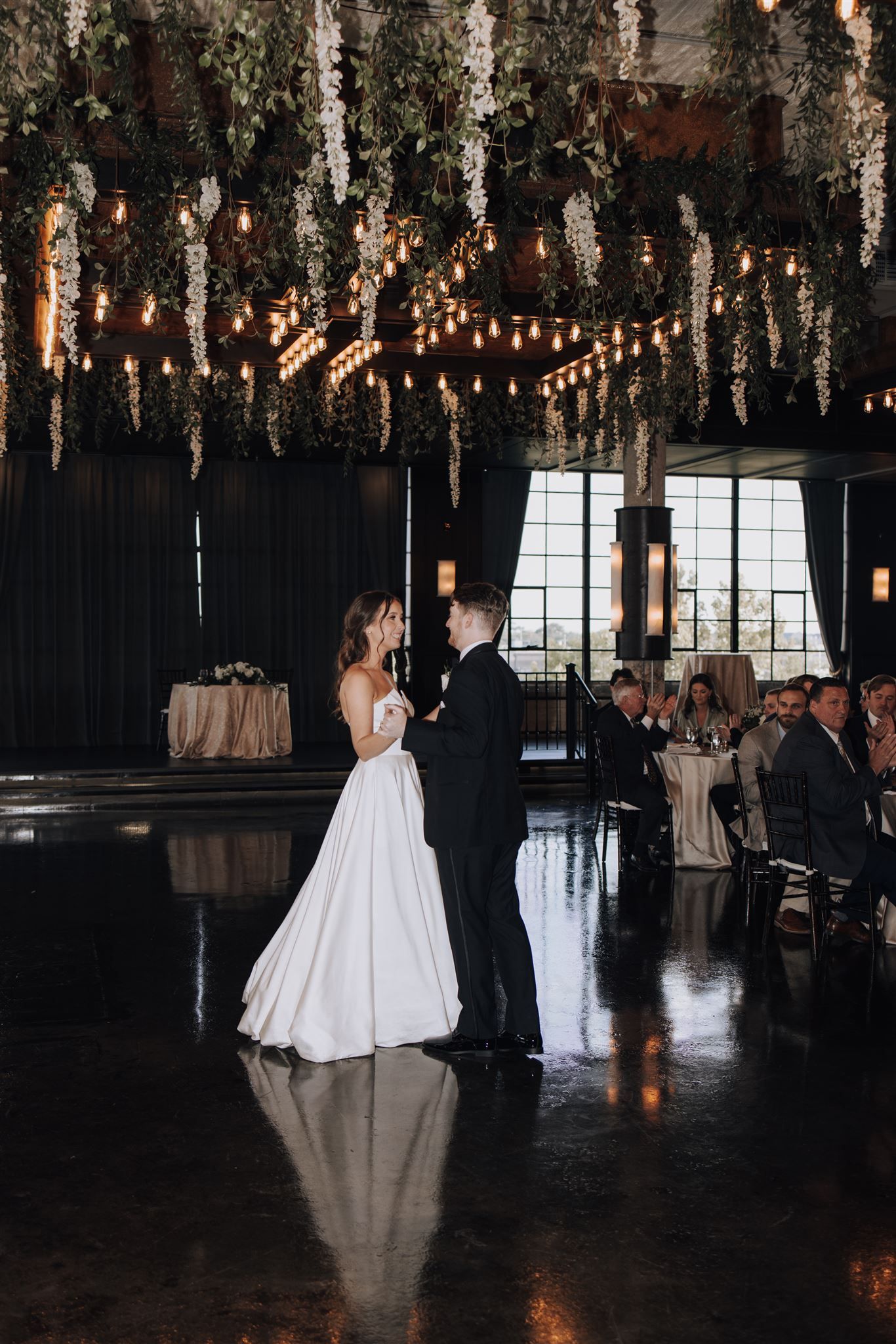 Couple shares first dance under hanging floral decor and string lights at wedding reception.