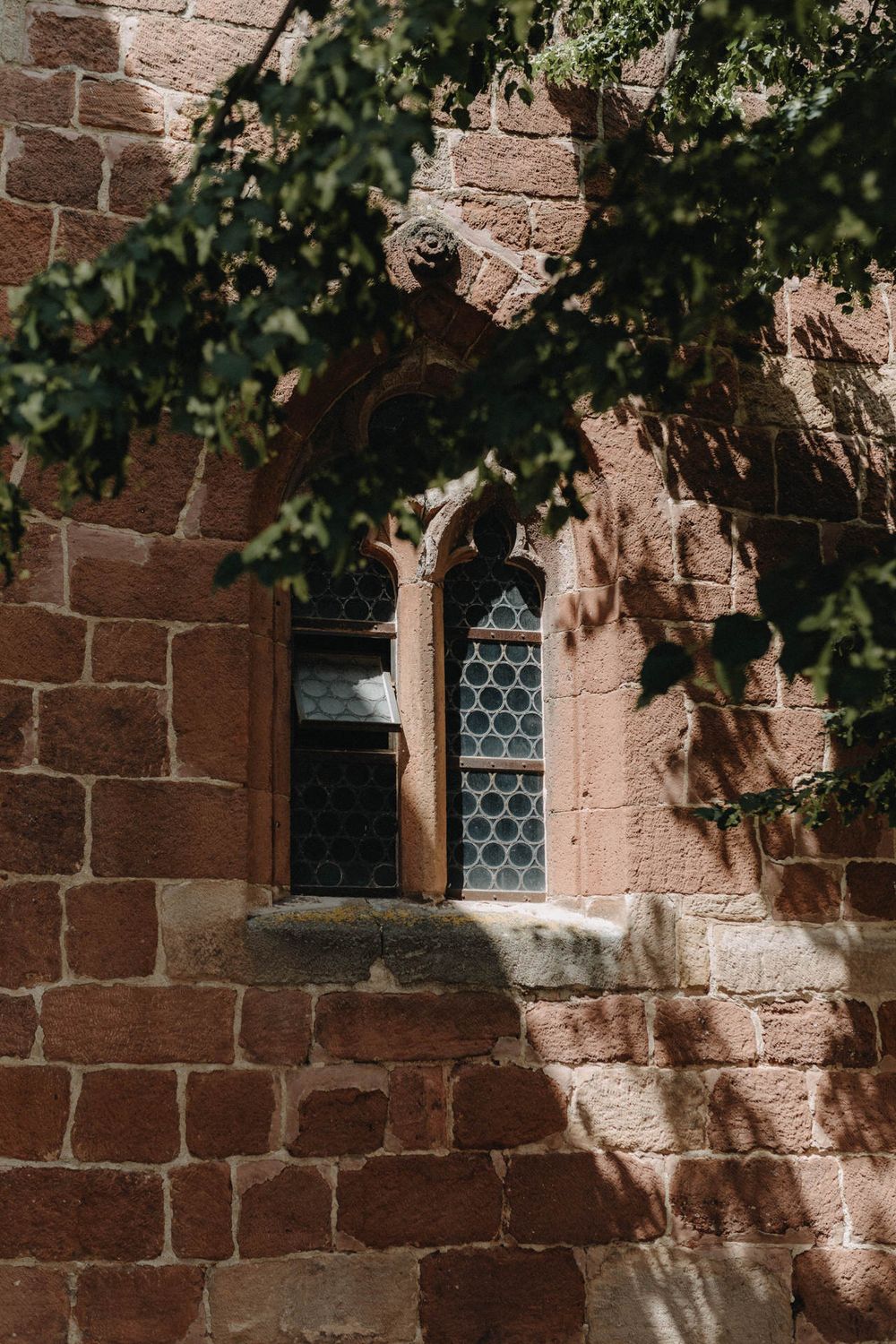 Gothic church window with stone archway framed by red brick walls and dappled sunlight.