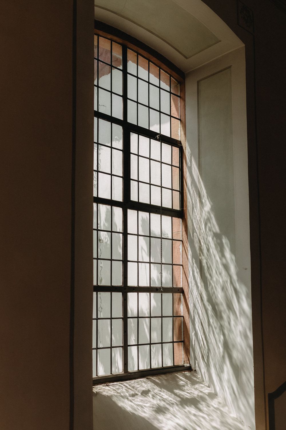 Tall arched church window with geometric panes casting dramatic shadows through sunlight.