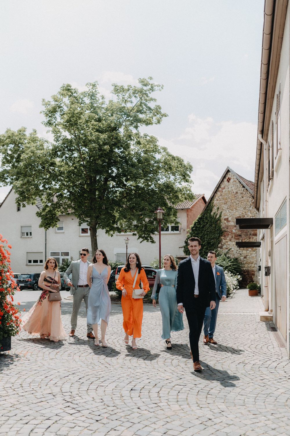 Wedding party walks down a cobblestone street wearing colorful formal attire on a sunny day.