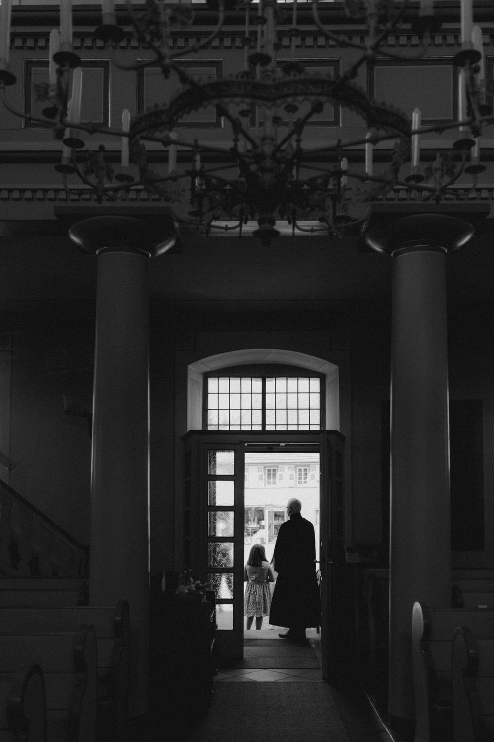 Silhouetted figures stand in a dramatic black and white doorway with architectural columns.