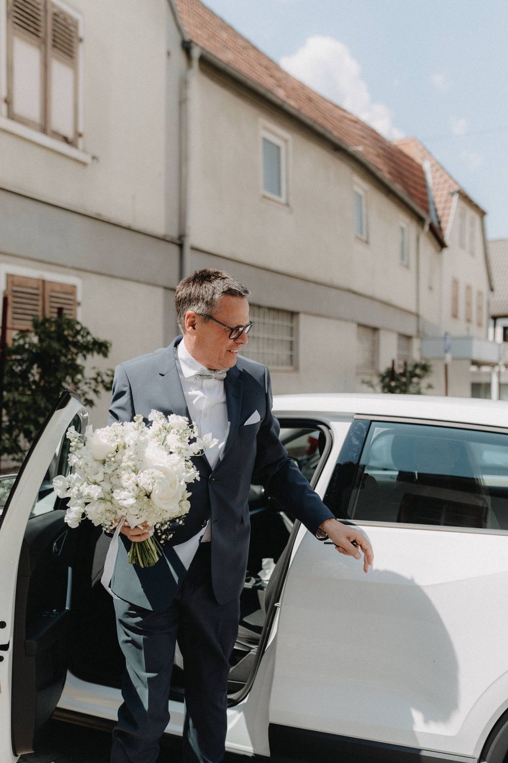 A formal-dressed person exits a white car holding an elegant white flower bouquet.