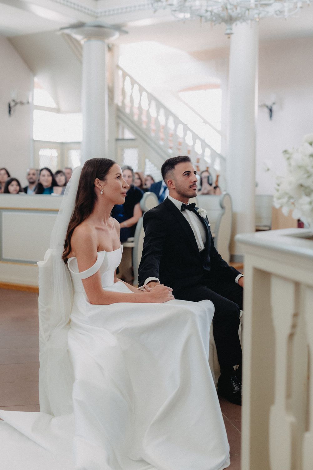 A bride and groom seated during their wedding ceremony in a traditional church setting with white columns.