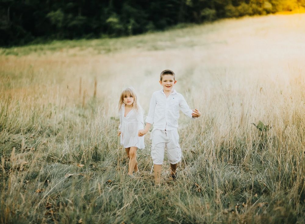 Two children holding hands while walking through a sunlit field of tall grass.