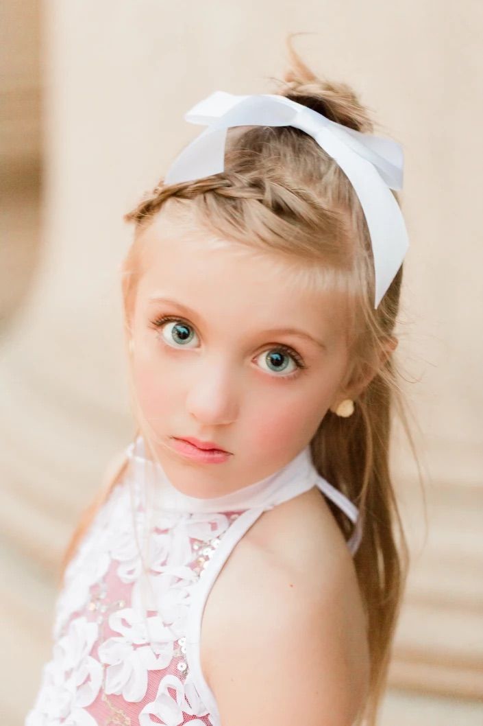 Portrait of a young performer in white headband and lace dress against a neutral background.