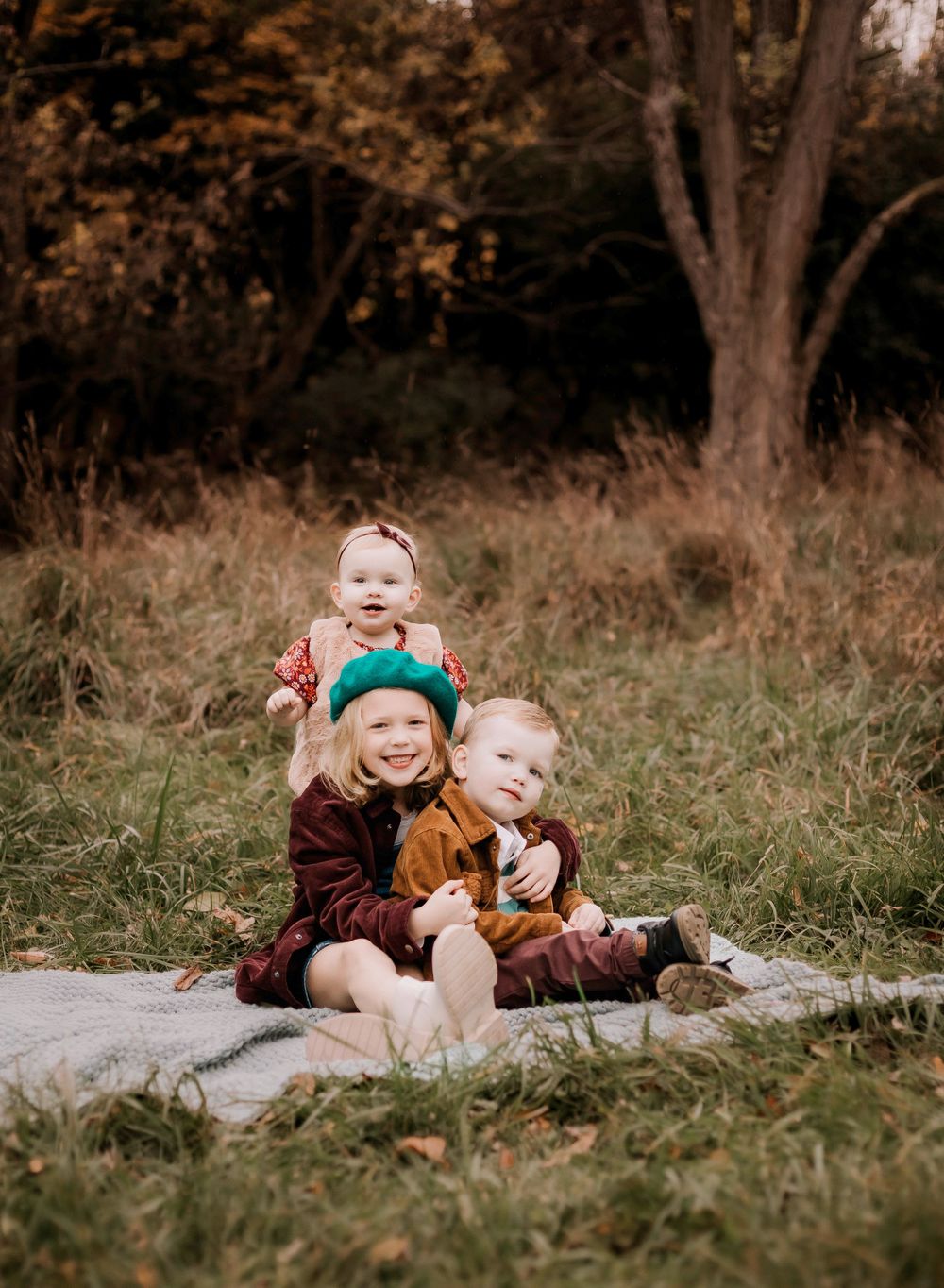 Three children sitting together and laughing on blanket in outdoor fall setting.
