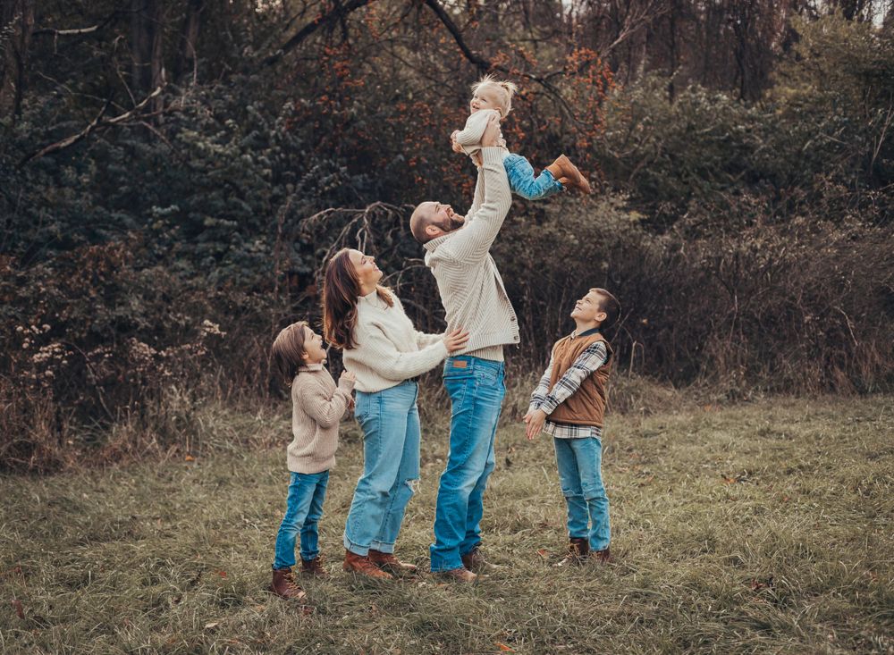 A family plays together in a field with a child being lifted playfully into the air.