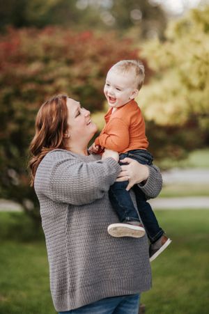 Parent in grey sweater lifting and playing with toddler wearing orange against autumn background.